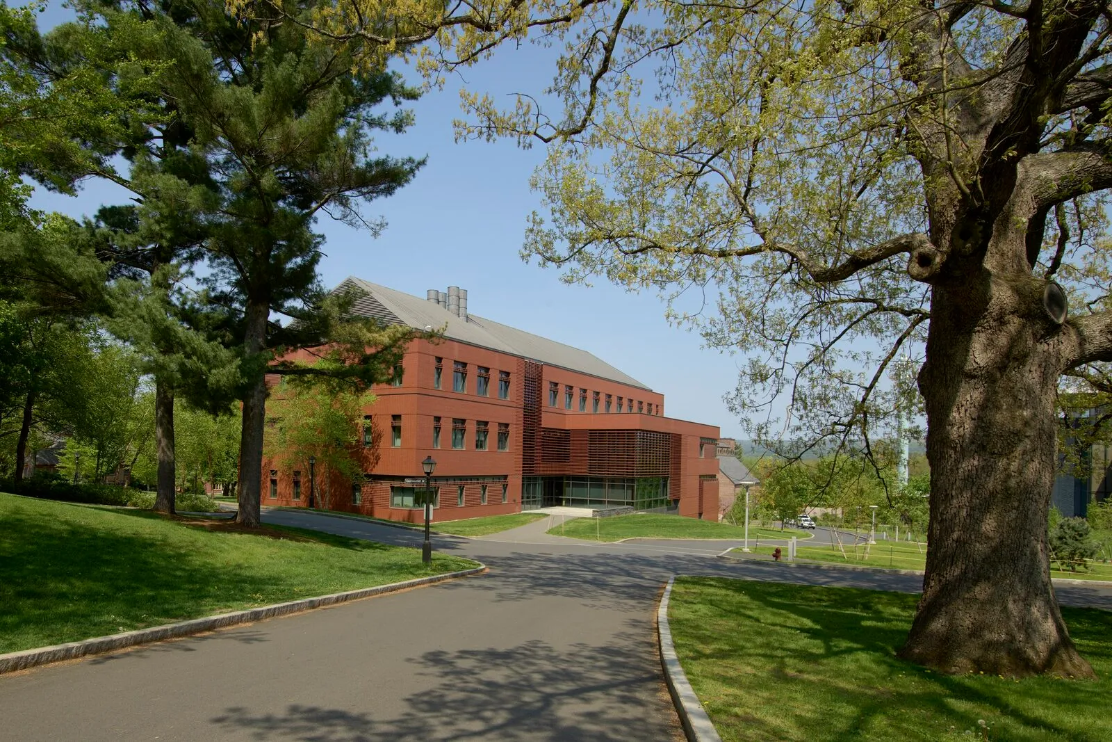 The Beneski Earth Sciences Building at Amherst College on a sunny late-spring afternoon. A three-story modern red-brick building with a gray hipped-shingle roof and a tall central atrium, set into a green campus landscape with a paved walkway curving toward the entrance, mature deciduous trees framing the view on both sides under a clear blue sky.