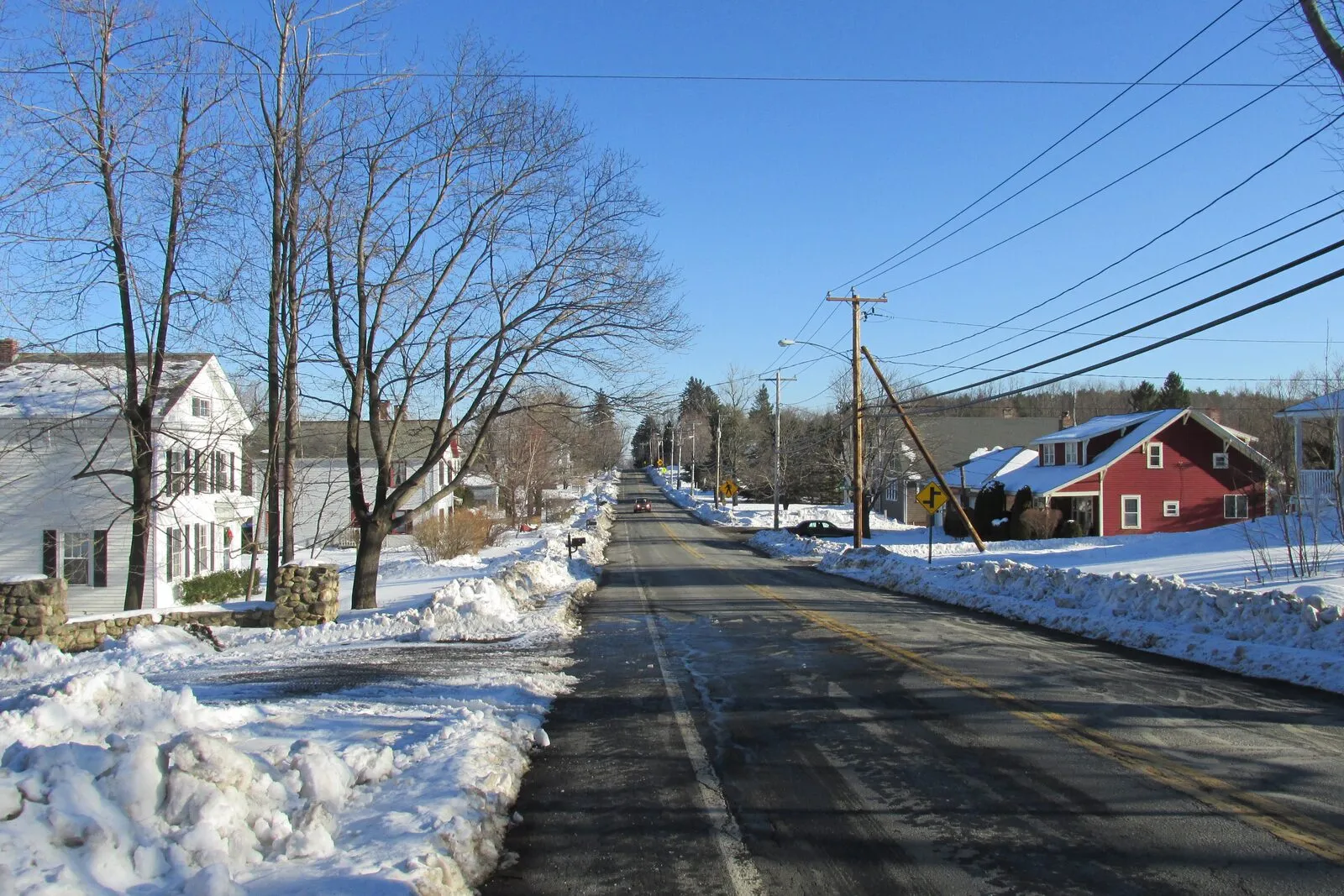 Looking down Main Street through the center of Blandford on a bright clear winter day: a wet and partly snow-covered two-lane road heading away into the distance with a long line of utility poles and overhead wires marching down the right side, packed snowbanks and a low fieldstone wall lining the left curb, a large white-clapboard Greek Revival house with green-shuttered windows behind bare deciduous trees on the left, a dark-red two-story barn-like commercial building with white trim and a snow-dusted roof on the right, more white New England houses receding behind it, low rolling wooded hills on the horizon under a deep cloudless blue sky.