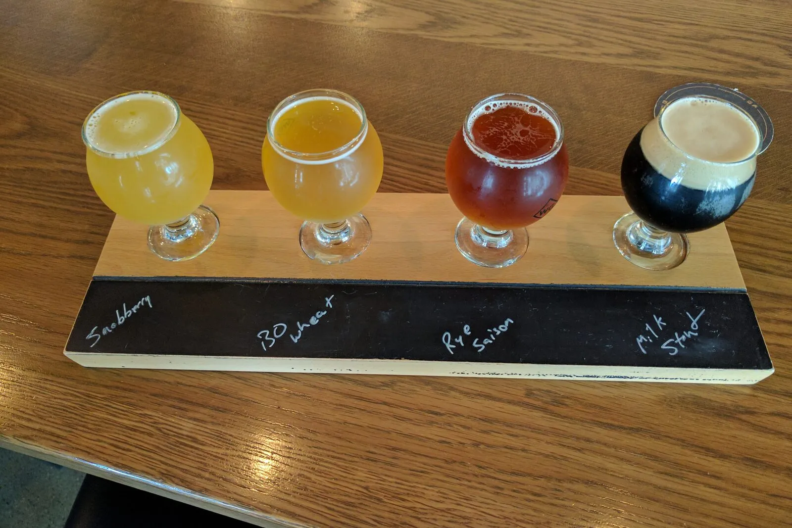 A wooden flight paddle on a bar table holding four small tulip glasses of beer — left to right a hazy pale gold, a clear straw-yellow, a deep amber-red, and a near-black stout topped with tan foam — each glass set into a circular cutout above a chalkboard strip with the four beer names written in white chalk. Indistinct dark-wood bar fixtures and a pale wall fade into the background behind the flight.