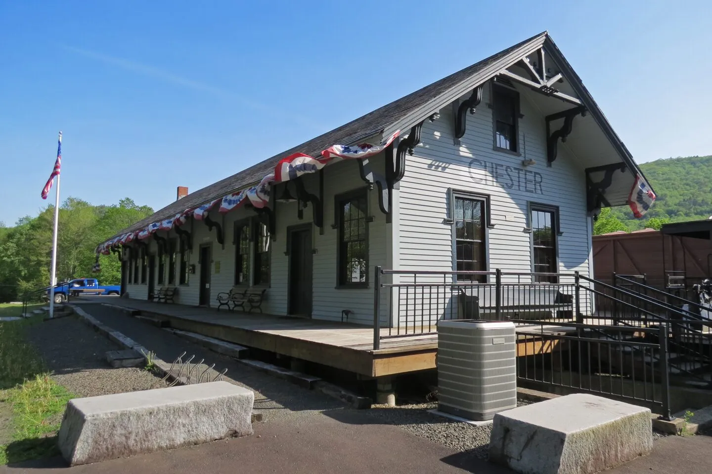 The 1841 Chester Railroad Station, a long, single-story white-clapboard depot with a steeply-pitched dark shingle gable roof, exposed wooden brackets, and the word CHESTER painted in dark letters on the gable end above paired stick-style brackets, on a clear sunny May morning. Patriotic red, white, and blue swag bunting hangs along the eaves of the trackside platform side, the station flag is up on a pole at the left, a wooden bench and a small water tank stand on the platform, a railroad freight car sits to the right, and a wooded green hillside rises behind under a deep blue sky.