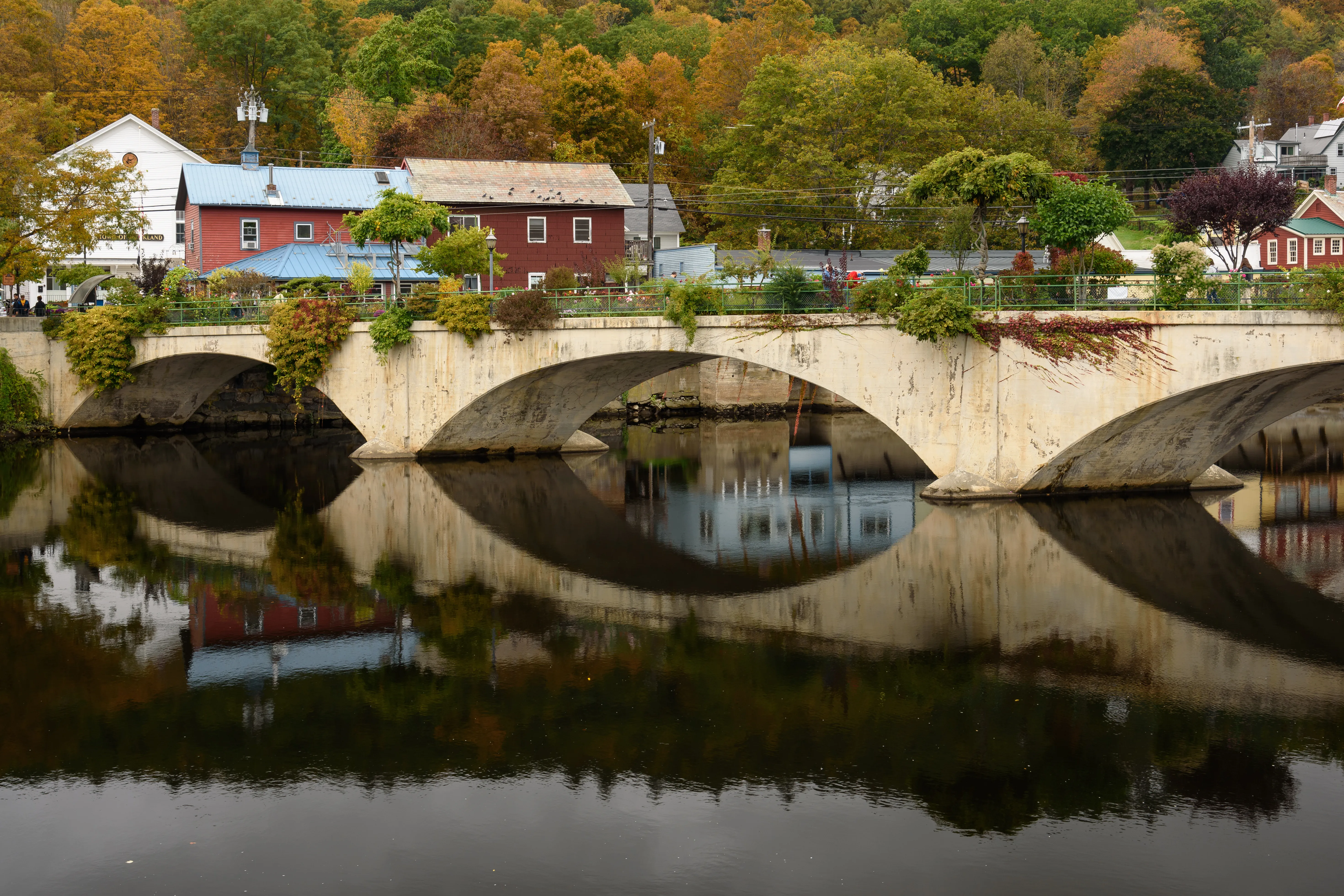 A stone arch bridge in Greenfield with peak fall foliage on the surrounding hills, reflected in the still water below.