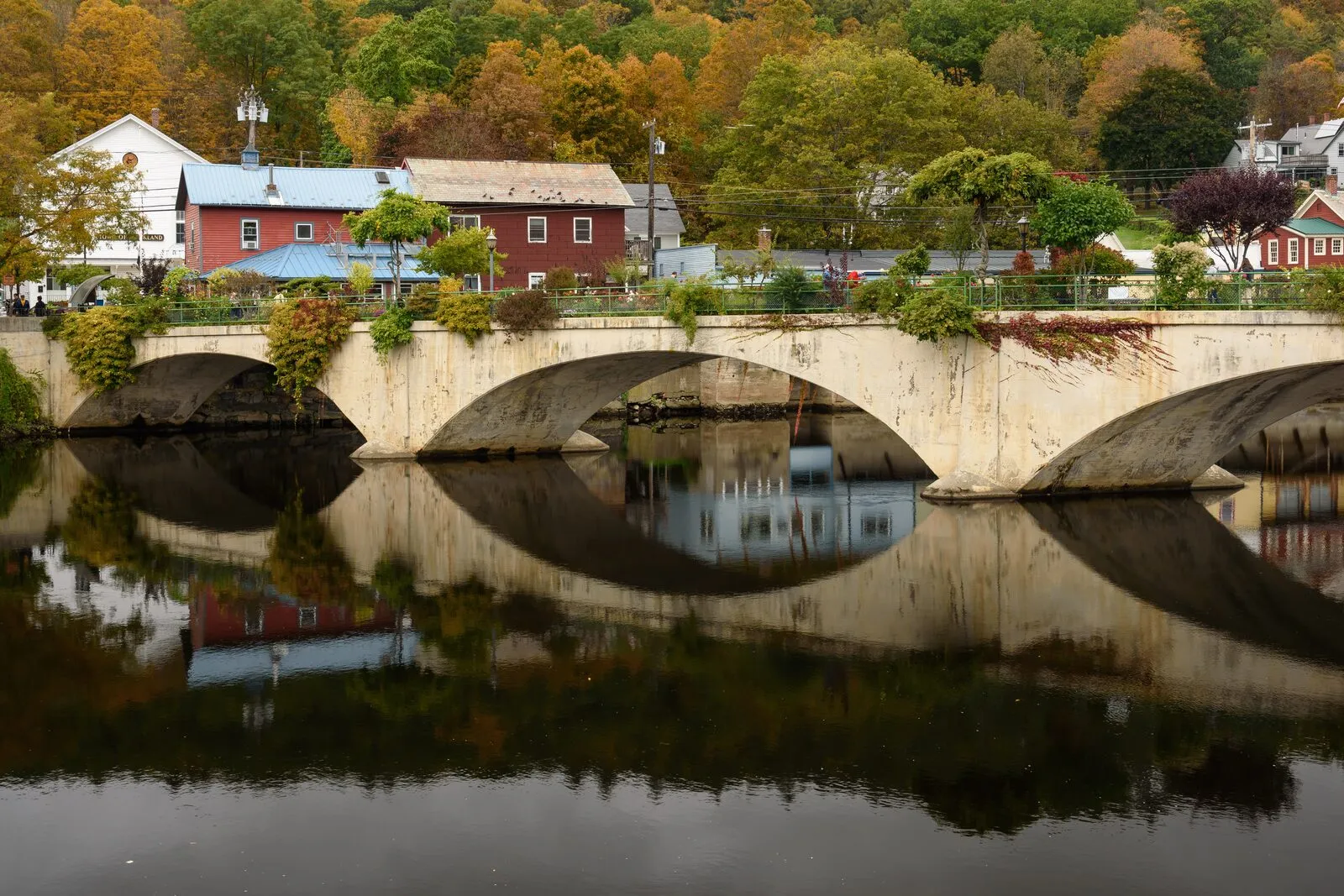 The stone-arch Bridge of Flowers in Shelburne Falls seen from the Deerfield River on an overcast October afternoon — vegetation cascading over the parapet, clean mirror reflections of the arches in the still water, red clapboard village buildings and a white spire behind the bridge, and a hillside of orange and yellow fall foliage beyond.