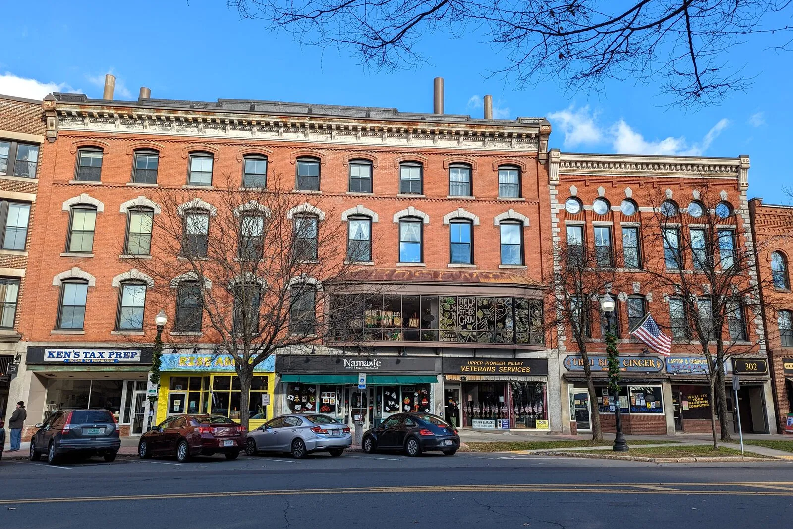 A row of four-story red-brick Italianate commercial blocks on Greenfield's Main Street. Ground-floor storefronts (Ken's Tax Prep, a Nepalese restaurant, Veterans Services) below tall arched and rectangular windows, bare winter branches reaching in from the right, parked cars along the curb, and a clear blue sky with thin clouds behind.