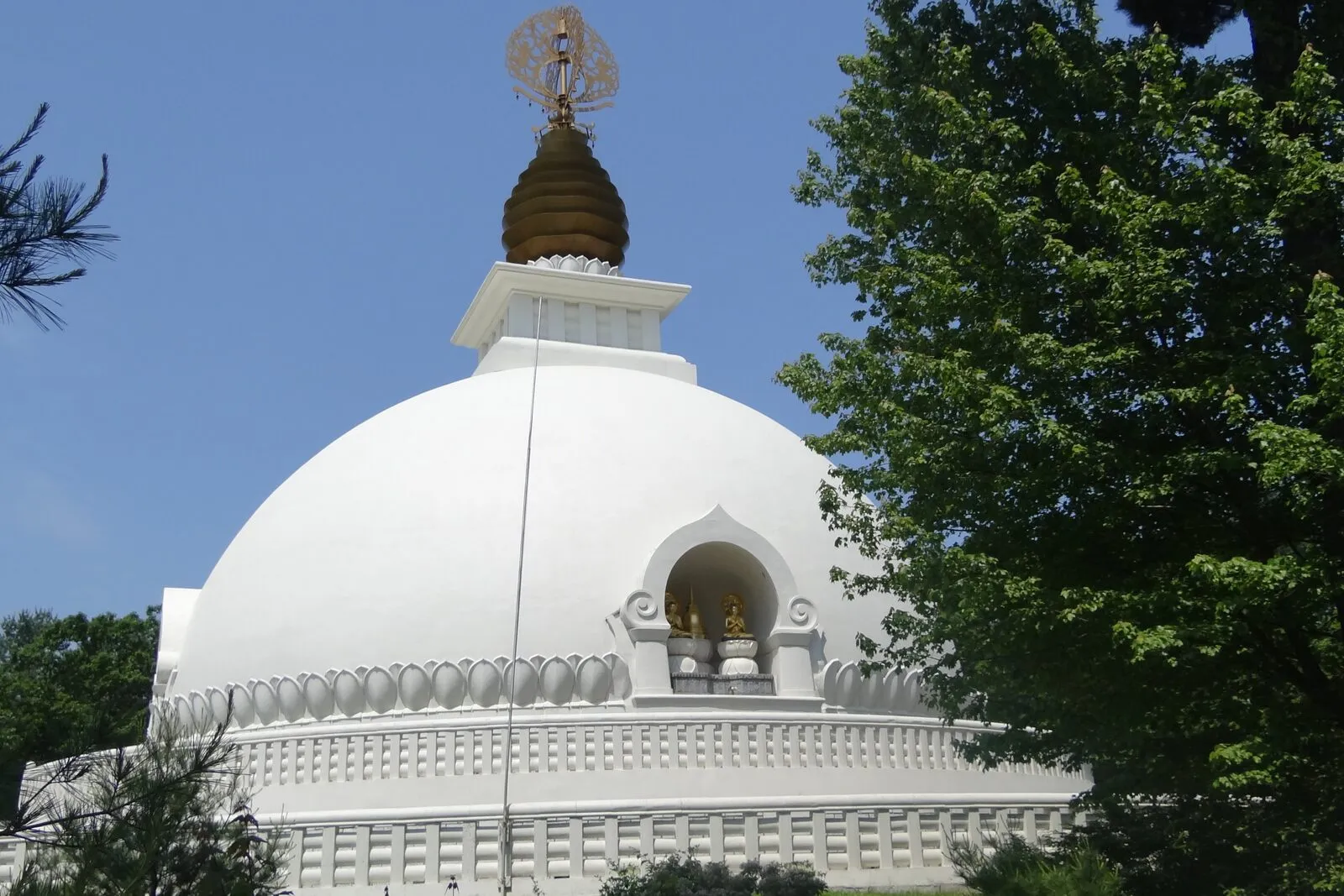 The New England Peace Pagoda on its hilltop in north Leverett on a clear summer day — a tall white-domed Buddhist stupa rising in two main tiers, the broad lower drum stepped back to a smaller upper drum capped by a tapering spire with a series of stacked gold-colored discs and a finial on top, golden Buddha figures set into recessed alcoves around the upper level facing the four cardinal directions, a wide white circular plaza of fitted stone paving running around the base, low stone walls and steps stepping down the hillside in the foreground, mature deciduous trees in full green leaf framing the structure to either side, and a deep blue sky behind.