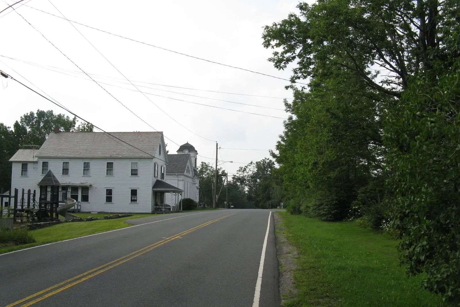 Middlefield Center on an overcast late-summer afternoon. A quiet two-lane country road with a double yellow center line curving gently into the distance, a large two-and-a-half-story white-clapboard house with dark roof and small covered porch on the left side of the road, a smaller white-clapboard church-like building with a square belfry tower visible just behind it, utility poles and overhead wires along the road's edge, a lone car parked further down the lane, a deep green hedgerow of mature deciduous trees in full leaf running the full length of the right side of the road, mowed grass shoulders on both sides, and a flat overcast gray sky overhead.