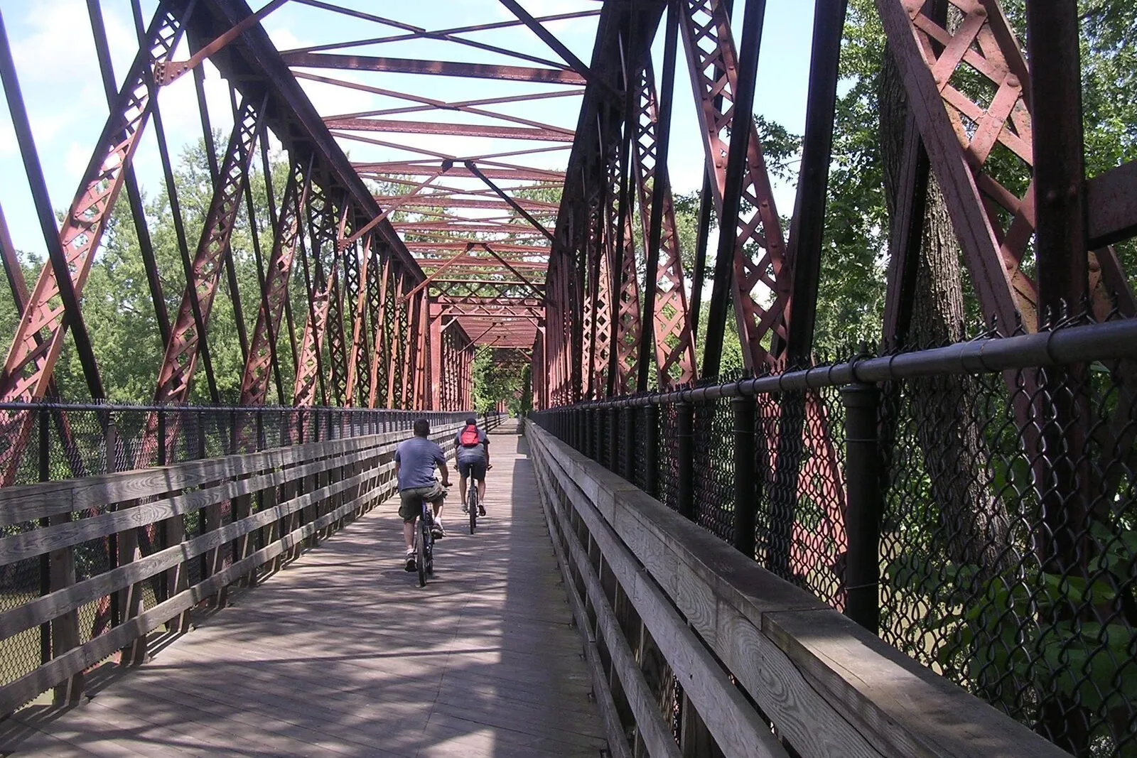 Two cyclists ride away from the camera across a long, rust-red iron truss bridge converted from a former railroad span. Diagonal lattice girders frame the view overhead and along both sides; a wooden plank deck and chain-link railings line the path. Summer foliage fills the gaps between the trusses on a bright, partly cloudy day.