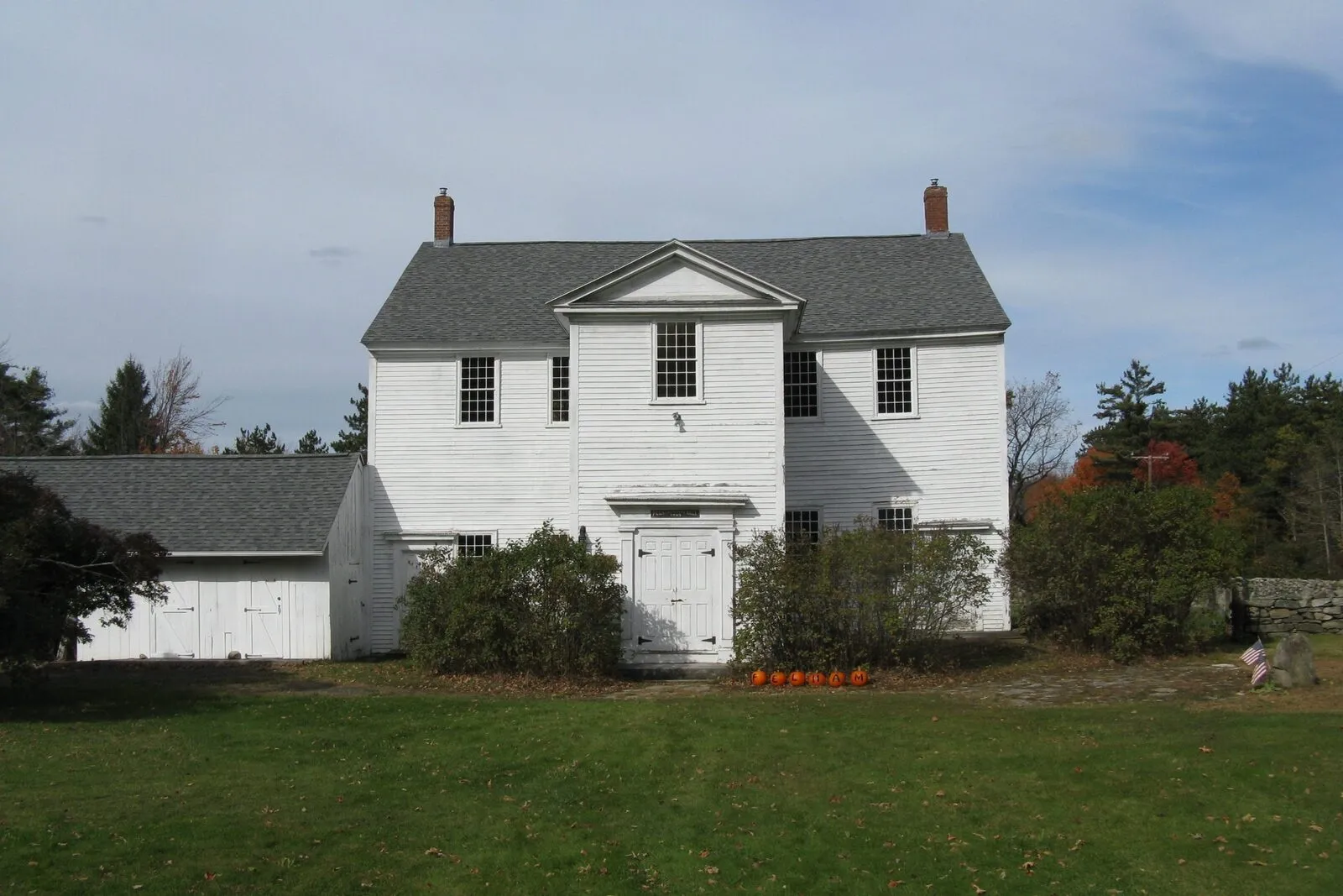 The Old Pelham Town Hall on a clear October afternoon: a tall two-story white-clapboard meeting house with a side-gabled gray-shingled roof, two narrow red-brick chimneys at the gable ends, a low central pediment over a single white double-leaf entry door, paired six-over-six sash windows flanking the entry on the ground floor and across the second story, a tall arched second-story window above the door with simple molded trim, a low one-story shed wing attached on the left with its own door, a low fieldstone wall running off to the right with a row of small American flags, a row of orange pumpkins along the base of the entry steps, a wide neatly mown lawn in the foreground, and a backdrop of dark conifers and just-turning sugar maples under a pale blue sky streaked with thin clouds.