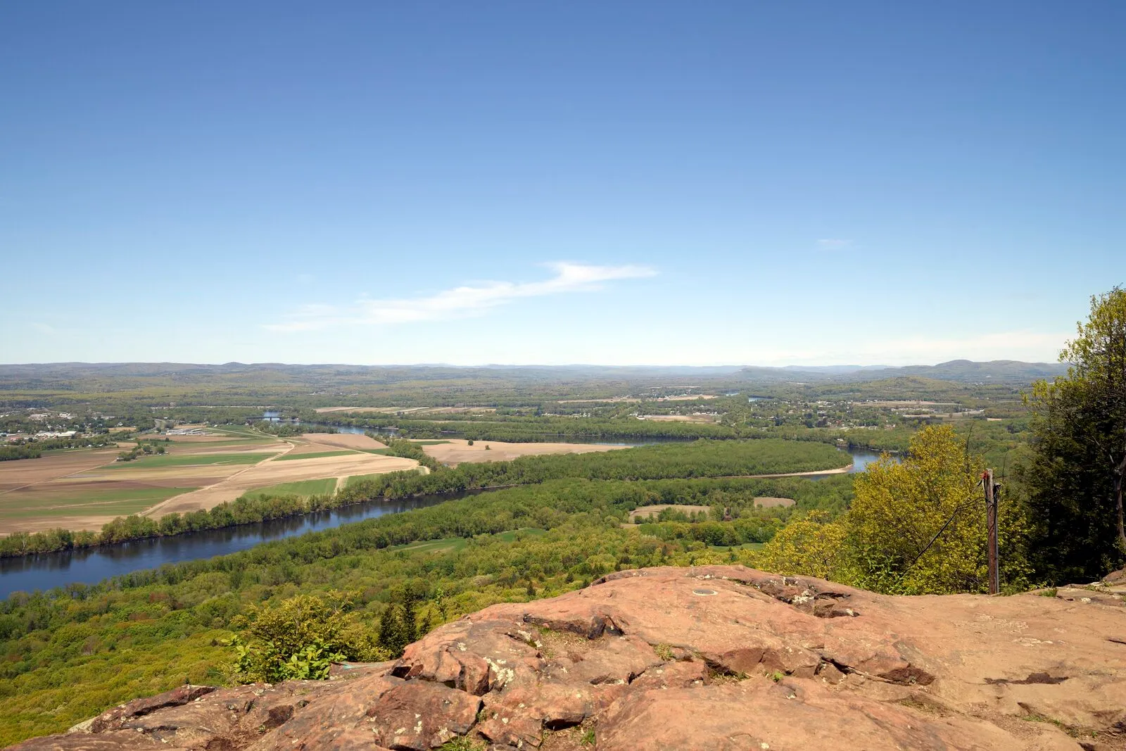 View south from the summit of Mount Holyoke in Skinner State Park, looking down across the Connecticut River as it curves through farm fields and wooded hills of the Pioneer Valley on a clear spring day, with red sandstone bedrock in the foreground.