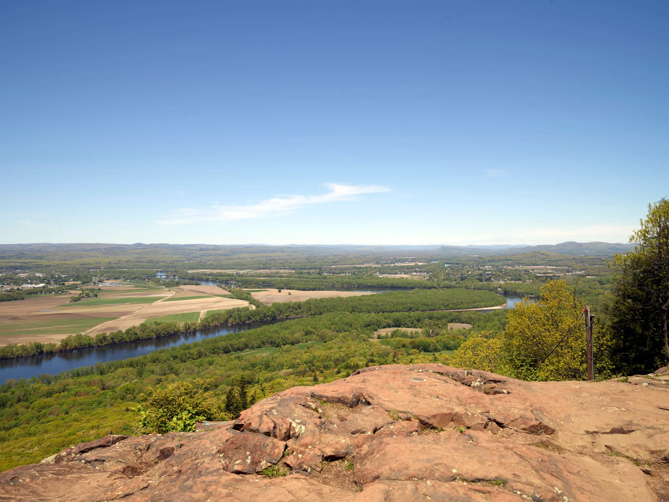 View south from the summit of Mount Holyoke in Skinner State Park, looking down across the Connecticut River as it curves through farm fields and wooded hills of the Pioneer Valley on a clear spring day, with red sandstone bedrock in the foreground.