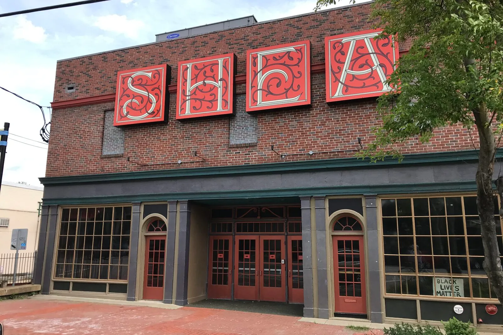 The Shea Theater facade at 71 Avenue A in Turners Falls on an overcast summer afternoon. A flat brick commercial-block front in red-brown brick with a horizontal row of three large red-bordered painted panels above the entry spelling 'SHEA' in ornate cream and gold scroll lettering, a continuous dark band running across the building below the signs, three sets of red-painted double entry doors set behind shallow arched openings flanked by square brick pillars, tall multi-pane storefront windows on either side, a small 'Black Lives Matter' sign in a window at the right, a leafy green street tree at the right edge, and a brick sidewalk in the foreground.