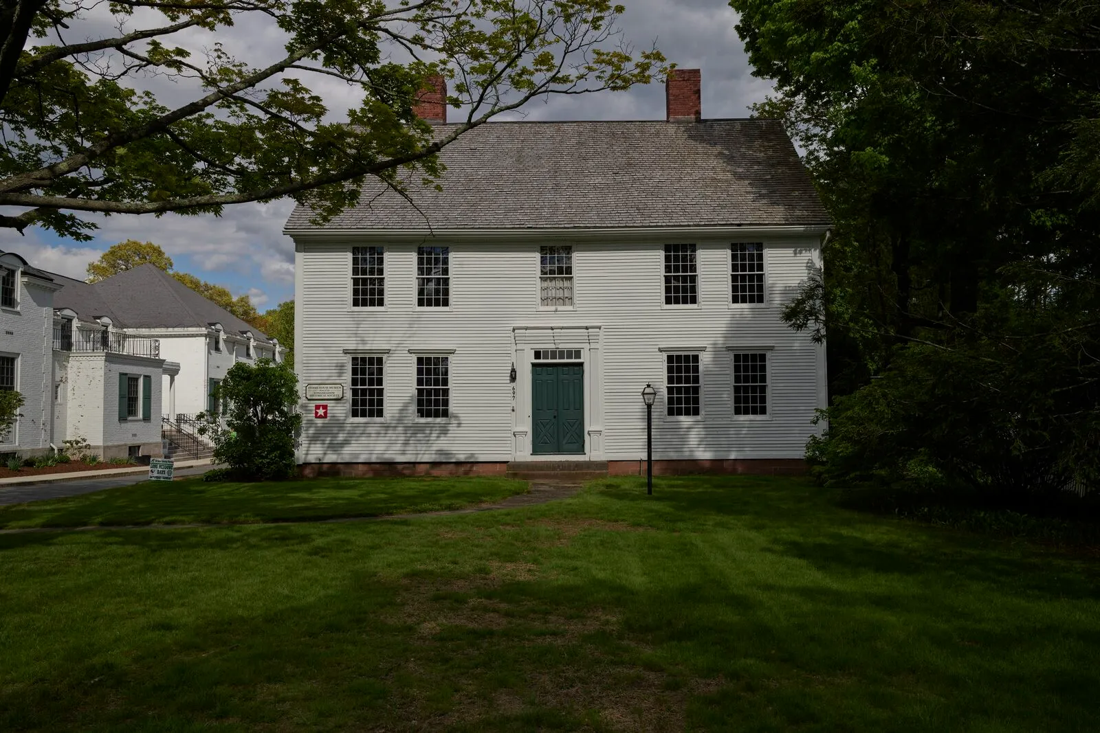 The Storrs House on Longmeadow Street, framed straight-on across a broad green lawn. A two-and-a-half-story Federal-era white-clapboard center-chimney colonial with a steeply pitched gray-shingled roof, two interior brick chimneys flanking the ridge, a five-bay symmetrical façade with twelve-over-twelve sash windows on both stories, a recessed center entry with a panelled green door, a black wrought-iron lamppost on the front lawn, mature deciduous trees in full leaf framing the house at left and right, a neighboring white-clapboard building partly visible on the left, and a sky of high broken cloud overhead.