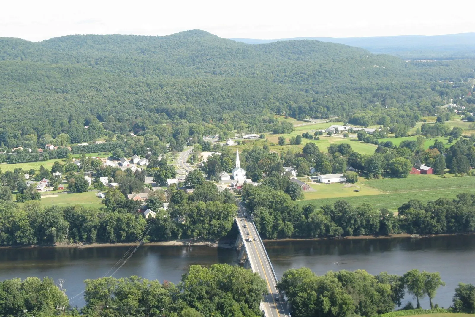 Sunderland village seen from the Mount Sugarloaf overlook on a sunny late-summer afternoon. The Connecticut River curves across the foreground with the green-painted steel Sunderland Bridge crossing it on Route 116, the village center clustered around the white spire of the First Congregational Church on the far bank, broad farm fields and tobacco barns spreading north and south, and the long forested ridge of Mount Toby filling the background under a clear blue sky with thin clouds.