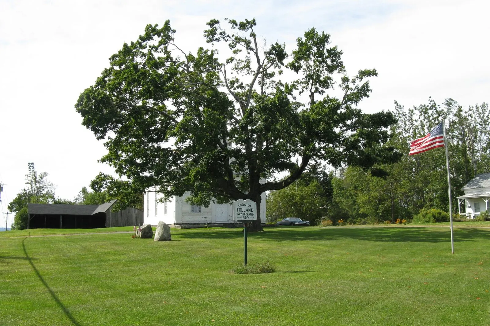 The tiny common at Tolland Center on a bright late-summer afternoon — a wide flat expanse of mown green lawn under a hazy pale sky, a single large mature deciduous tree spreading at the center with a small white-painted town sign on a post just below it, an American flag flying from a tall white flagpole at right, the long low form of a white-clapboard town building with a gable roof in the middle distance behind the tree, an open dark equipment shed at far left, a glimpse of another white New England house at right edge, and a continuous line of dark green woods closing off the back of the green.