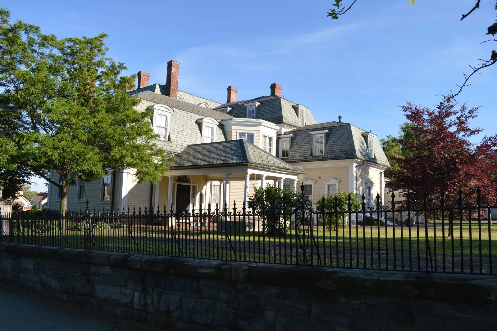 The pale-yellow clapboard Wistariahurst mansion seen from Cabot Street on a summer afternoon: a long gable-and-dormer slate roof with red-brick chimneys, a hipped-roof front portico over the main entry, a black cast-iron fence atop a random-ashlar stone wall running across the frontage, a deep-green leafy tree on the left, and a red-leaf copper-beech on the right under a clear blue sky.